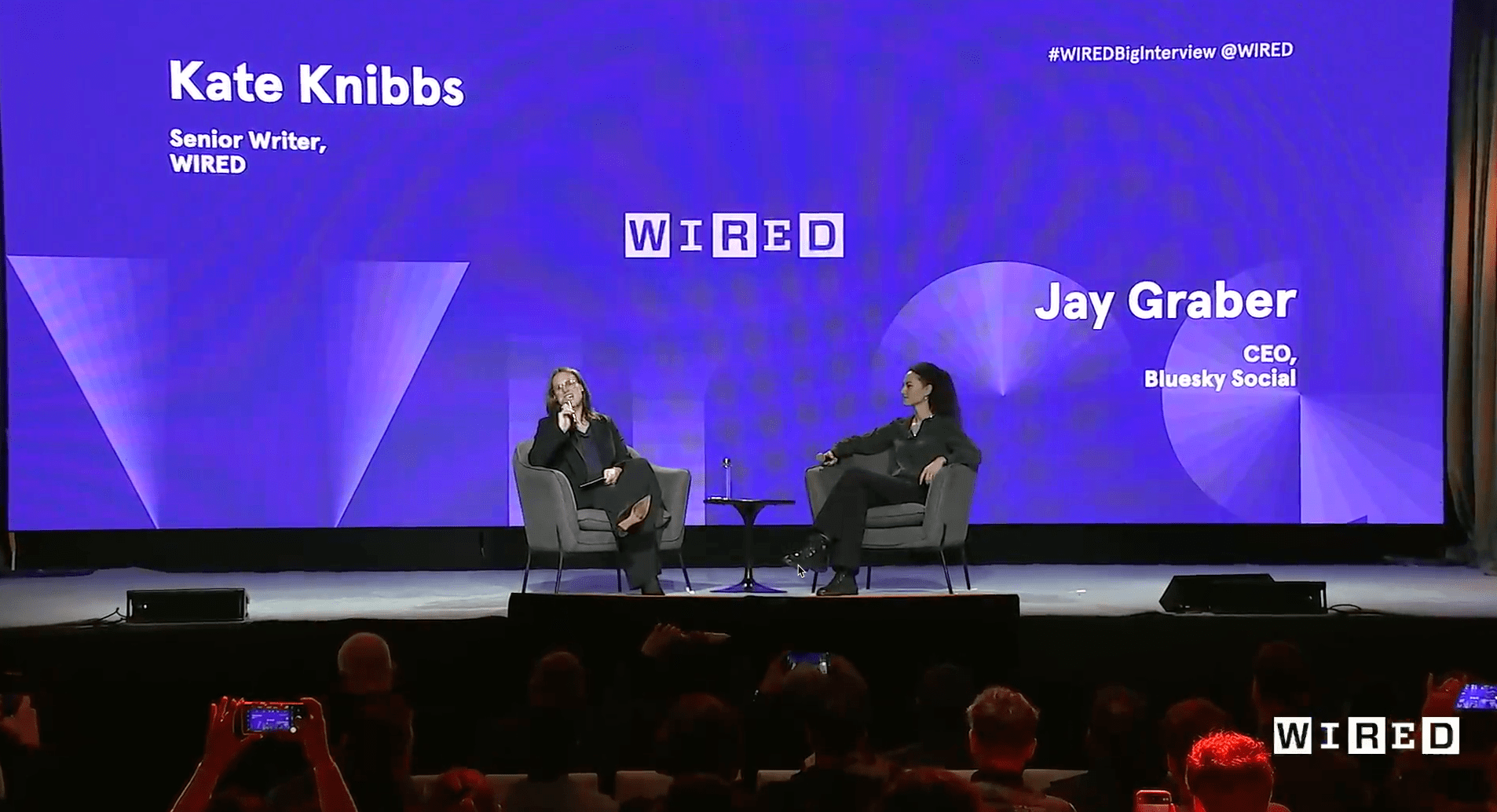 The image shows a stage where two people are seated on armchairs, engaged in a conversation. The background is a bright purple screen displaying text. On the left side, it says:

"Kate Knibbs Senior Writer, WIRED"

On the right side, it reads:

"Jay Graber CEO, Bluesky Social"

The top-right corner includes the hashtag: "#WIREDBigInterview @WIRED." Both individuals are holding microphones, and they appear to be part of a live interview or discussion, likely organized by WIRED. The stage has a small table with a bottle of water between them, and there are audience members visible in the foreground.