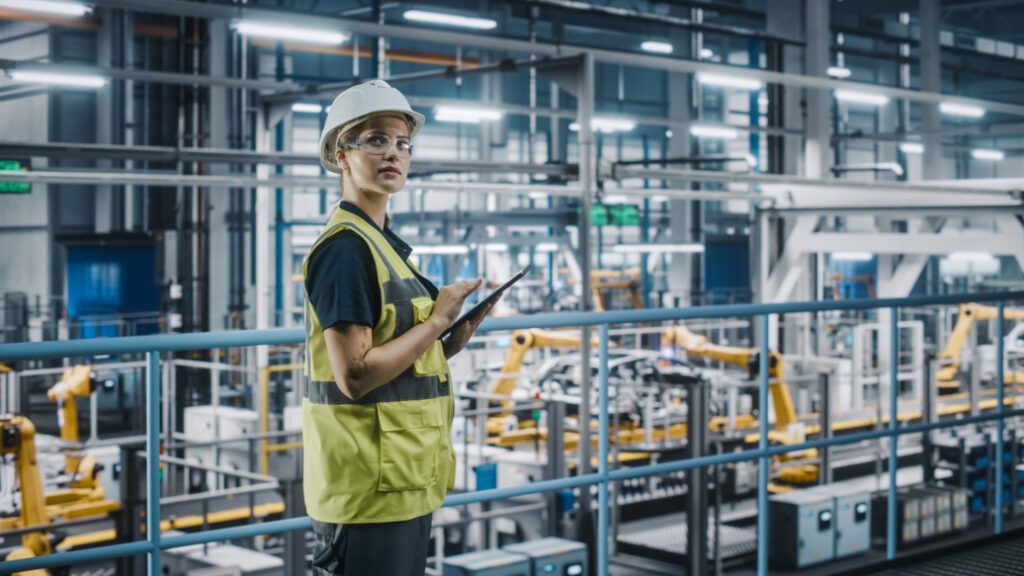 Female Car Factory Engineer in High Visibility Vest Using Laptop Computer. Automotive Industrial Manufacturing Facility Working on Vehicle Production with Robotic Arms. Automated Assembly Plant.