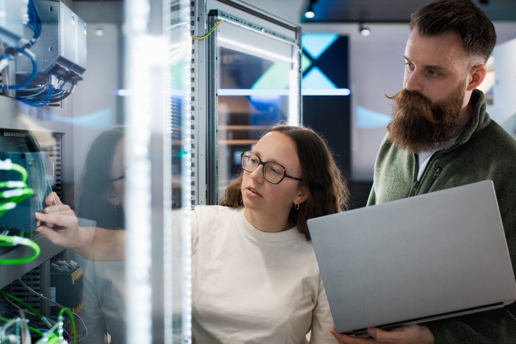 Two automation engineers inspecting the programmable logic controllers on the shop floor while holding a laptop for referencing